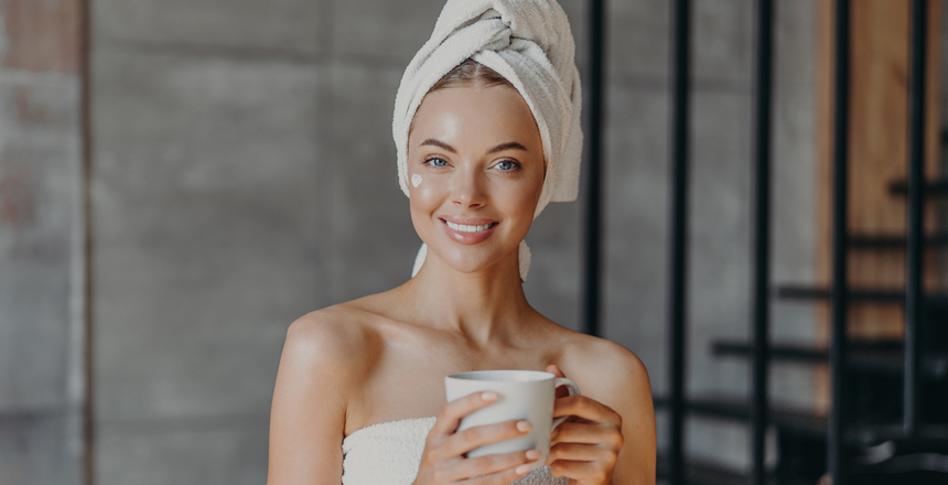 Attractive smiling young woman with makeup, toothy smile, poses bare shoulders, wrapped in towel, drinks hot beverage stands indoor has cheerful expression applies moisturizing cream on face