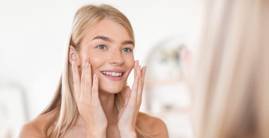 A young blonde woman with a radiant complexion is gently touching her cheeks and smiling, standing in a bright, airy bathroom. She appears to be engaging in a skincare routine, possibly after a Fire & Ice Facial treatment.